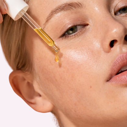 Close-up of a woman applying a dropper of yellow facial glow oil to her face.
